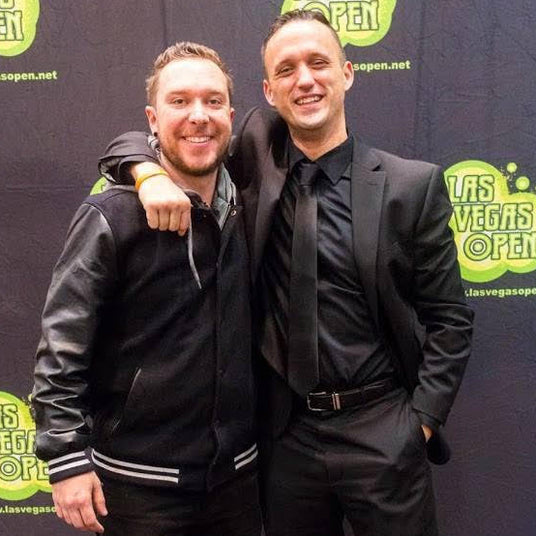 Two men posing together in front of a Las Vegas Open event backdrop. The man on the left wears a black leather jacket, black pants, and black sneakers.