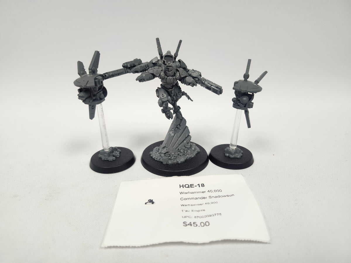 Unpainted gray assembled Warhammer 40,000 miniature set on three black round bases: a central armored figure on a sculpted rocky base flanked by two small drone units on clear flying stands. A label card in front reads "HQE-18", "Warhammer 40,000 Commander Shadowsun" and "Tau Empire".