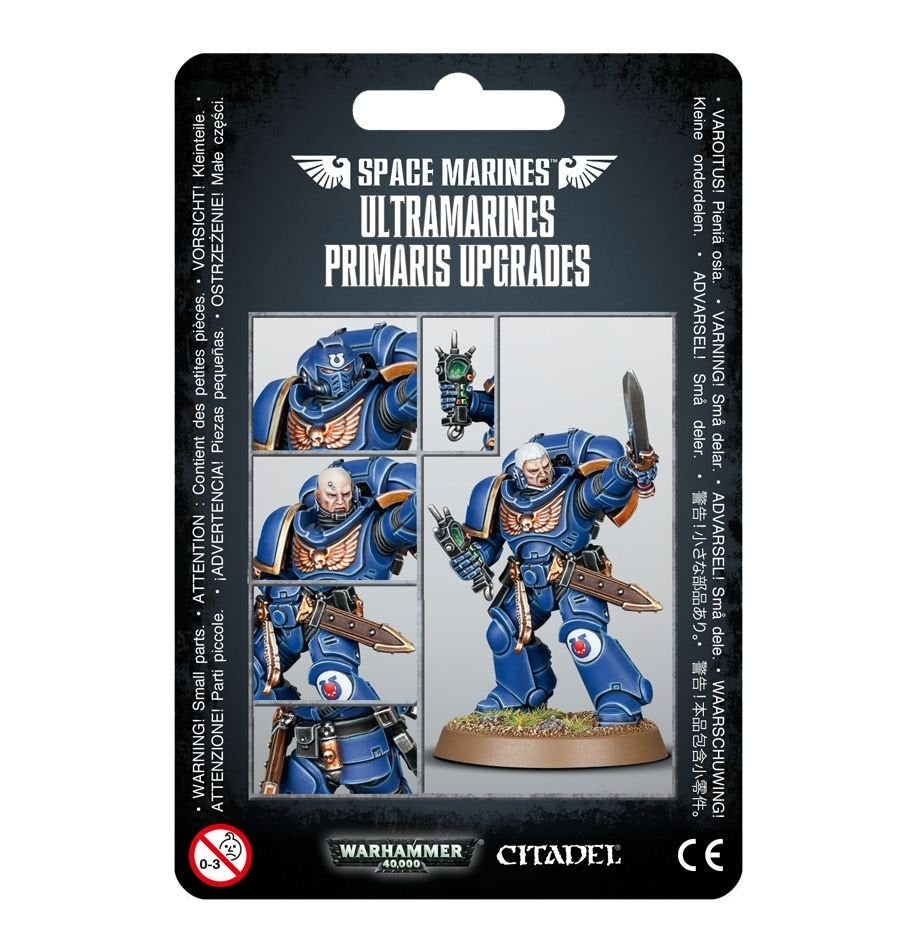 Front of a black Ultramarines upgrade package, labeled “SPACE MARINES” and “ULTRAMARINES PRIMARIS UPGRADES.” It shows a blue-armored Space Marine holding a sword and pistol, with smaller close-ups of the helmet, head, weapon and armor details.