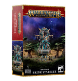 Boxed Seraphon Skink Starseer miniature, shown on the front and left side of a red-and-gold package. The artwork depicts a turquoise, ornamented lizard-like figure on a raised platform against a starry forest; the box reads “WARHAMMER AGE OF SIGMAR” and “SKINK STARSEER.”