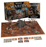 Warcry: Nightmare Quest game set displayed with an illustrated brown-and-blue box and a “MIGHT AND MADNESS” booklet behind a brown play mat, miniature figures, ruined stone scenery, bare trees, cards and reference sheets. The box reads “WARCRY” and “NIGHTMARE QUEST.”