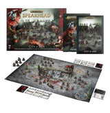 Warhammer Age of Sigmar Spearhead: City of Ash contents on white: a box cover with gold "SPEARHEAD" and "CITY OF ASH" lettering over a red-caped helmeted warrior and a miniature battle scene, two rulebook booklets, and a grey cobblestone game board set up with painted miniatures on round bases, terrain pieces and cards.