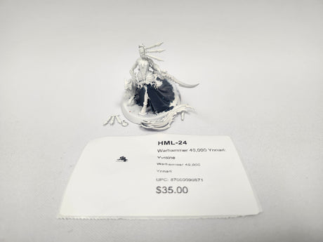 Unpainted white plastic miniature of Yvraine, a female figure with a feathered headdress, flowing gown with a dark lower skirt, and a curved sword, standing on a round base with loose sprue parts beside it on a plain light background. A white label card in front reads "HML-24" and "Warhammer 40,000 Ynnari: Yvraine".