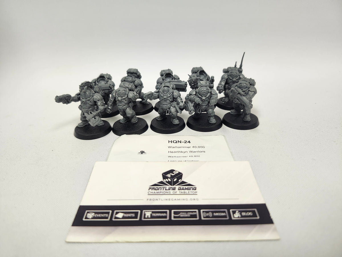 Ten unpainted grey Hearthkyn Warriors miniatures on round black bases, posed with weapons and backpacks, arranged behind two white cards. The top card reads "HQN-24 Warhammer 40,000 Hearthkyn Warriors Leagues of Votann"; the lower card shows a die logo with "FRONTLINE GAMING CHAMPIONS OF TABLETOP".