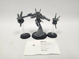 Unpainted gray assembled Warhammer 40,000 miniature set on three black round bases: a central armored figure on a sculpted rocky base flanked by two small drone units on clear flying stands. A label card in front reads "HQE-18", "Warhammer 40,000 Commander Shadowsun" and "Tau Empire".