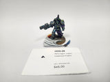 Painted miniature of an Alpha Legion Cataphractii Praetor, a heavily armored warrior in teal and blue-green power armor with a purple cloak, holding a large gun, on a round base with a sculpted white banner and a small blue blade. A white label card in front reads "HOG-28", "Alpha Legion -Legion" and "Cataphractii Praetor".