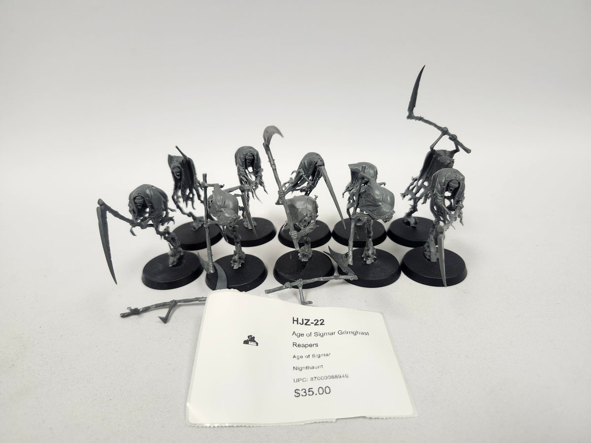 Group of unpainted gray plastic Age of Sigmar Grimghast Reaper miniatures on round black bases, hooded skeletal figures wielding scythes and polearms, with loose weapon parts on the table and a white label in front reading "HJZ-22 Age of Sigmar Grimghast Reapers" and "Nighthaunt".