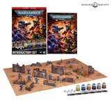 Warhammer 40,000 Introductory Set contents: a box cover reading "START HERE", "WARHAMMER 40,000" and "INTRODUCTORY SET", a matching handbook, and a laid-out battle scene with blue Space Marine and green Ork miniatures among cardboard ruin terrain on a sandy mat, plus paint pots, dice and a ruler. "WARHAMMER COMMUNITY" logo appears top right.