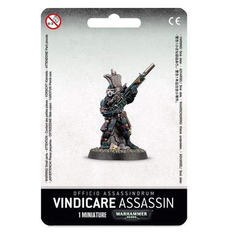 A Vindicare Assassin miniature is shown on its round scenic base, wearing dark teal and black armor and aiming a long rifle upward, on white-and-gray card packaging labeled “VINDICARE ASSASSIN” and “1 MINIATURE.”