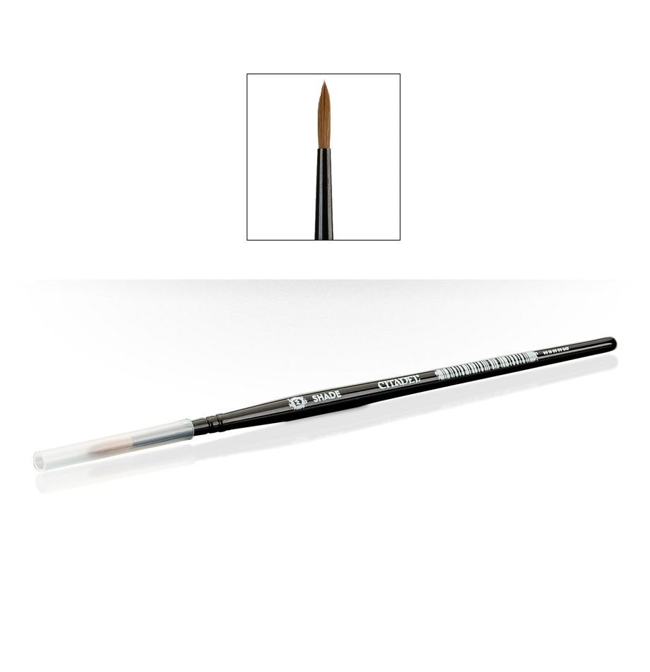A slim black Citadel shade brush lies diagonally with a clear protective cap over one end and pointed brown bristles at the other. An inset shows the brush tip close up; the handle reads “SHADE” and “CITADEL.”
