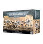 A rectangular Warhammer 40,000 box shows white and gray Crisis Battlesuit miniatures with small drones on rocky terrain against a tan backdrop. The front reads “WARHAMMER 40,000,” “T’AU EMPIRE,” “CRISIS BATTLESUITS” and “9 MINIATURES”; the right side shows additional miniature artwork.