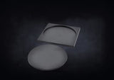 Two dark gray miniature base pieces are shown on a dark background: a flat round base in front and a square carrier behind it with a large circular recessed opening.