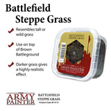 A clear plastic tub of dark green and brown steppe grass flock is shown at an angle beside the headline “Battlefield Steppe Grass.” Its olive and red label includes a grass sample window and “The Army Painter” logo.