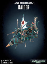 A dark, angular Drukhari Raider miniature with teal edging, a white sail bearing a red emblem, gold accents, and small crew figures, shown against a dark background. The text reads “DRUKHARI,” “RAIDER,” and “WARHAMMER 40,000.”