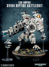 A white XV104 Riptide Battlesuit miniature with red markings stands on a rocky, grass-tufted base, flanked by two small drones on clear stands. The graphic reads “TAU EMPIRE XV104 RIPTIDE BATTLESUIT” and “3 MINIATURES.”
