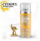 A yellow-orange Citadel Zandri Dust spray can with a clear plastic cap stands beside a small tan-painted armored miniature holding a raised weapon. The can reads “CITADEL,” “MODEL PAINT,” and “ZANDRI DUST.”