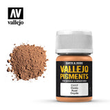 A small bottle of rust-colored pigment with a white ribbed cap and black-and-orange label stands beside a mound of brown powder. The label reads “VALLEJO PIGMENTS” and “Óxido Rust Oxyde.”