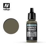 A small olive-drab Vallejo primer bottle with a tall black dropper cap stands beside a round swatch of matching paint. Its black label reads “SURFACE PRIMER” and “US Olive Drab”; the Vallejo logo appears above.