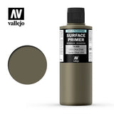 A tall olive-drab Vallejo primer bottle with a black cap and black-and-white label stands beside a matching round color swatch. The label reads “SURFACE PRIMER” and “USA Olive Drab,” with the “vallejo” logo above.