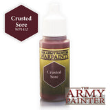 A small dropper bottle of dark reddish-brown paint with a white cap and green-and-yellow label reading “THE ARMY PAINTER WARPAINTS” and “Crusted Sore.” A matching color swatch reads “Crusted Sore WP1412.”